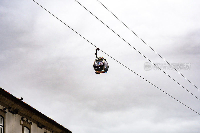 在多云的天空下，盖亚新城(Vila Nova de Gaia)附近的缆车的缆车舱悬挂在悬挂的缆索上，将游客从路易斯一世桥(Don Luis I bridge)运送到波尔图长廊(Porto prowalk)。